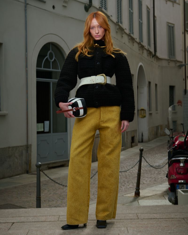 A fashionable red-haired woman poses in mustard-yellow pants and a black jacket, carrying a geometric bag, on a city street.