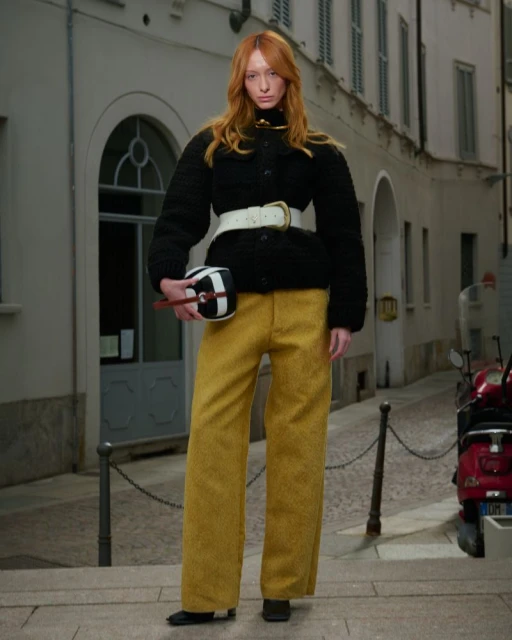 A fashionable red-haired woman poses in mustard-yellow pants and a black jacket, carrying a geometric bag, on a city street.