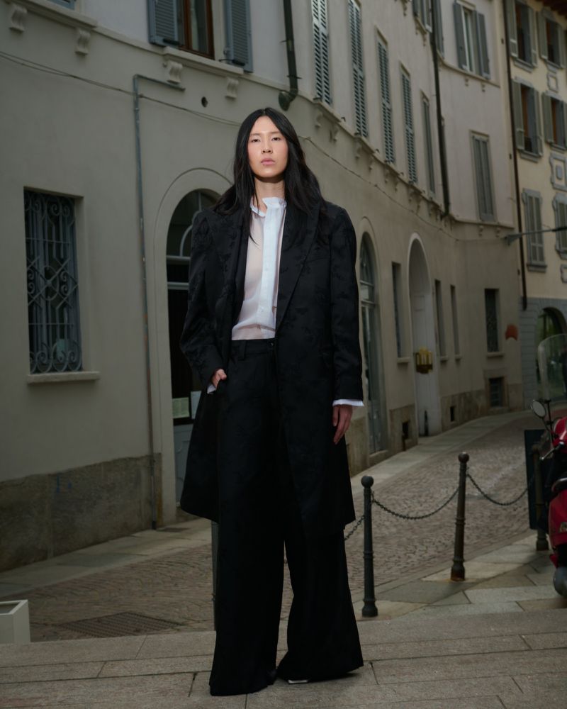 An Asian female fashion model in a black coat and wide-legged trousers stands on a narrow cobblestone street at dusk in an editorial shot.