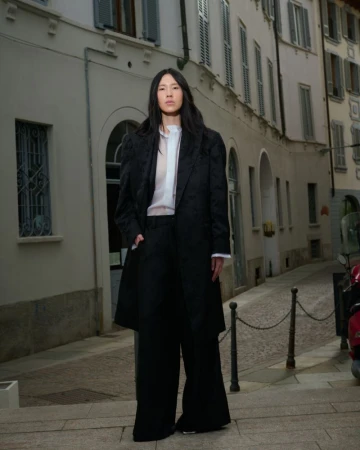 An Asian female fashion model in a black coat and wide-legged trousers stands on a narrow cobblestone street at dusk in an editorial shot.