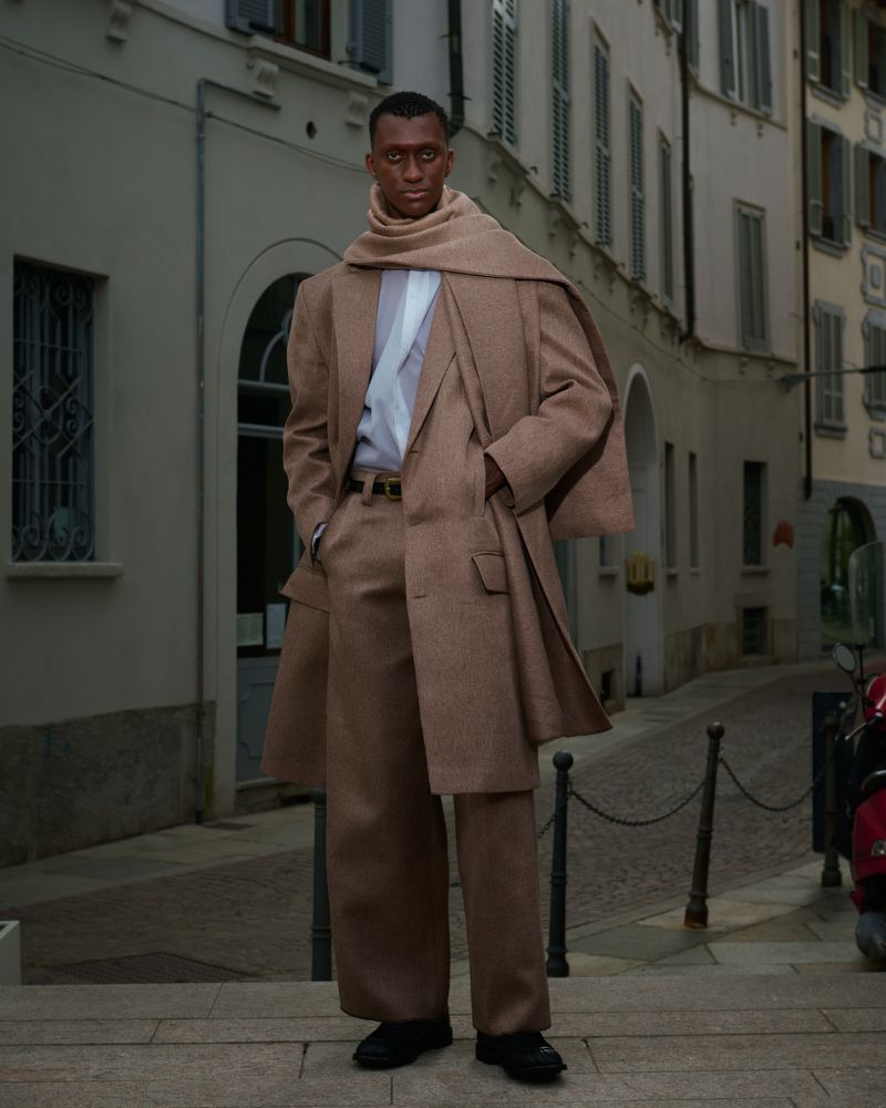 A dark-skinned man poses on a city street, wearing a tan overcoat and scarf, paired with wide-legged pants, in an urban environment.