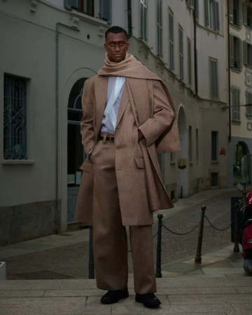 A dark-skinned man poses on a city street, wearing a tan overcoat and scarf, paired with wide-legged pants, in an urban environment.