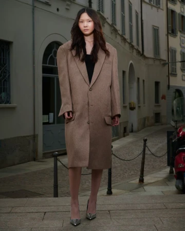 A stylish Asian woman models an oversized tan coat and silver heels on a cobblestone street in front of a beige urban building.