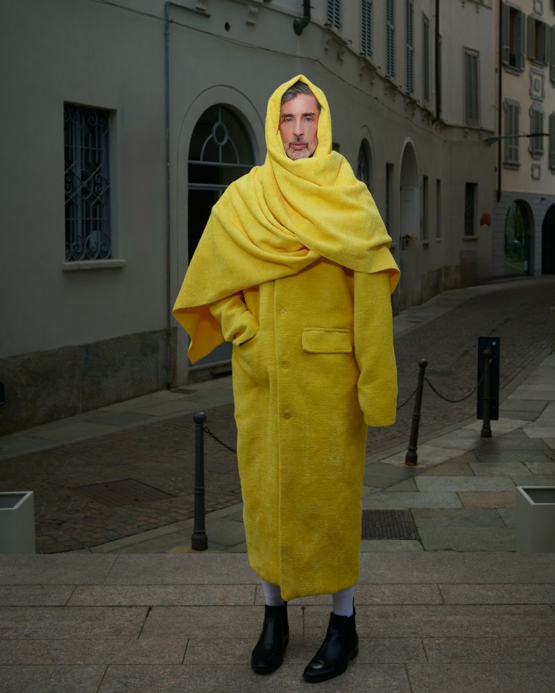 An adult stands outside on a cobblestone street wearing a long yellow coat with a scarf, a hood, and black boots.
