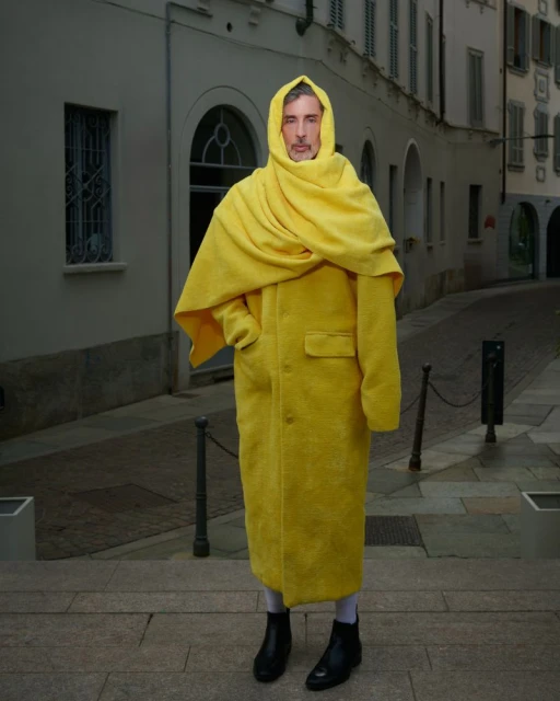 An adult stands outside on a cobblestone street wearing a long yellow coat with a scarf, a hood, and black boots.