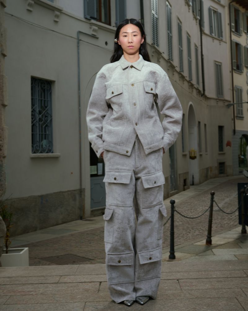 Asian woman in gray jacket and pants stands on stairs in front of buildings. Fashion shot with street photography aesthetic.