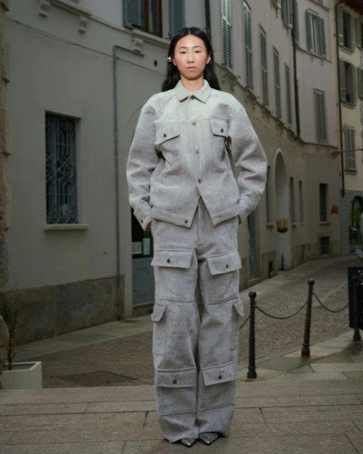 Asian woman in gray jacket and pants stands on stairs in front of buildings. Fashion shot with street photography aesthetic.