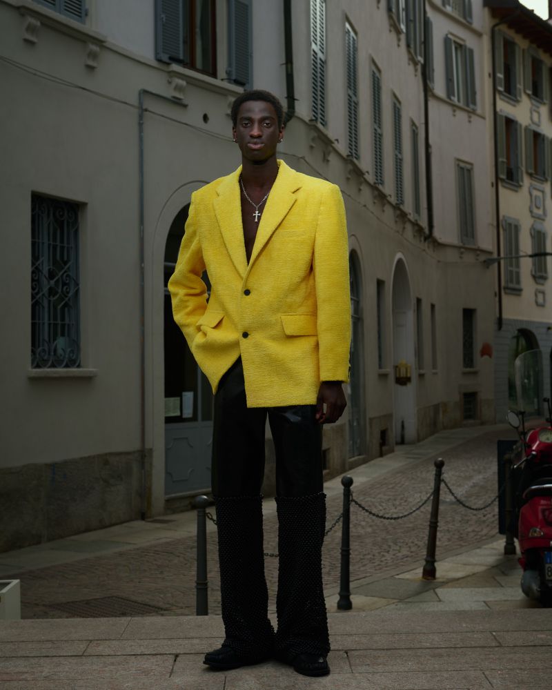 A young Black man in a yellow blazer and black pants stands on a city street with white buildings and red scooter in the background.