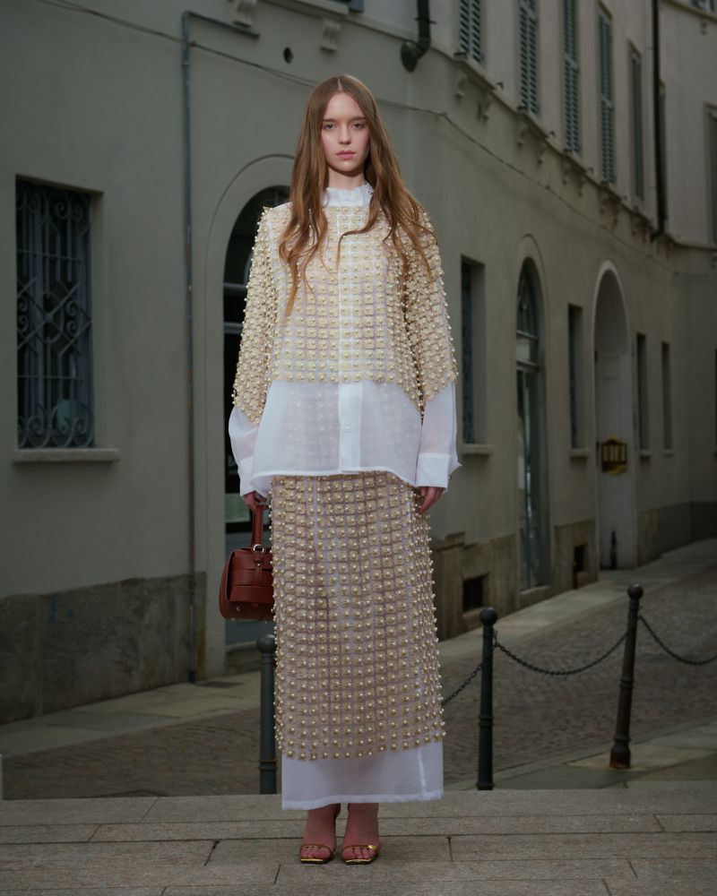 Full-shot of a young adult woman in a beaded top and skirt, with a brown handbag, standing on steps with a building in the background.