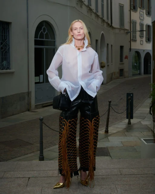 Fashionable woman in a sheer white blouse and leather pants with fringe posing on a city street with neutral building facades.