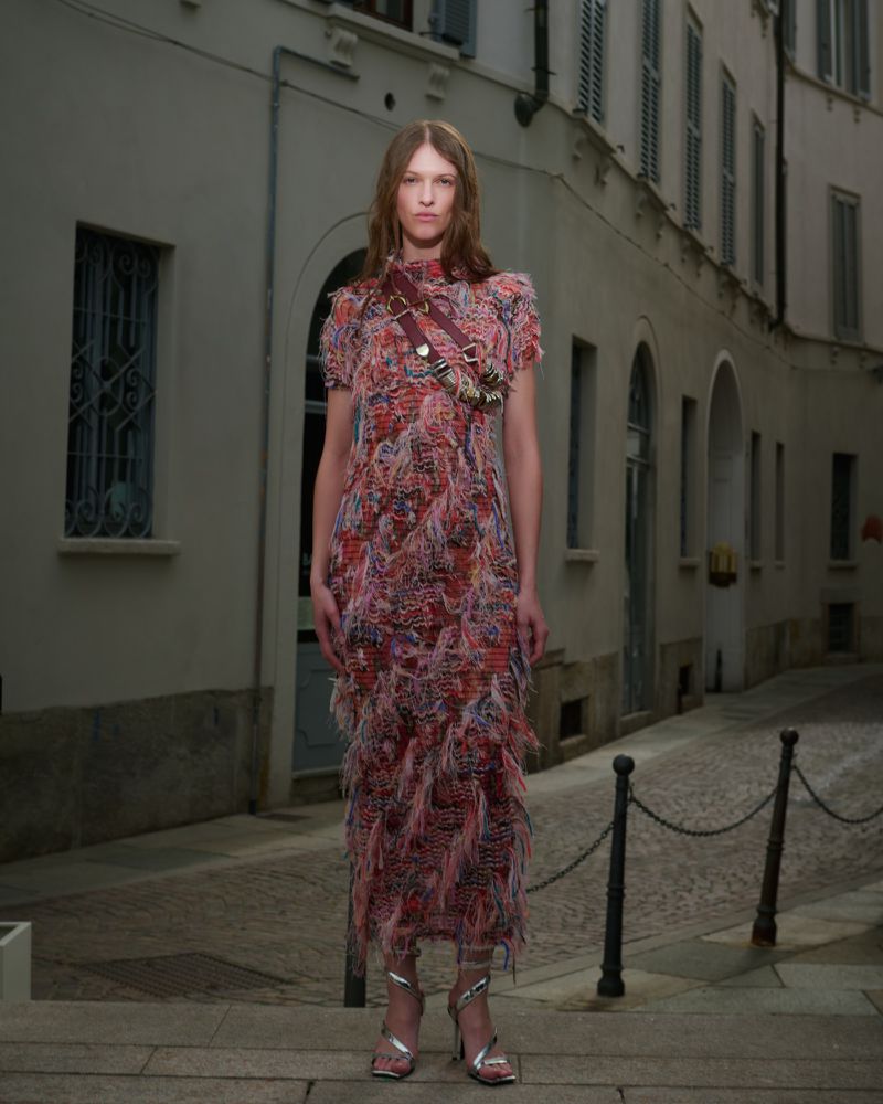 Woman in patterned dress and silver heels stands on cobblestone street in Milan, Italy, with historic buildings in the background.