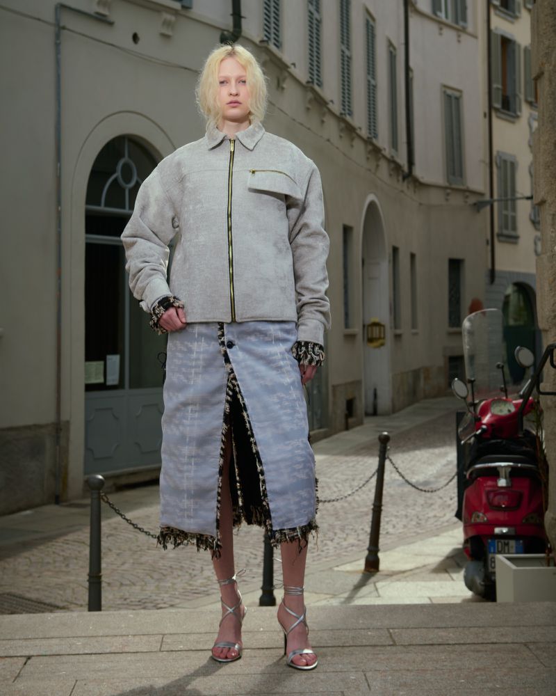 A fashion shot of a young adult woman in a grey jacket and light blue skirt, silver heels, standing on a street in an urban setting.