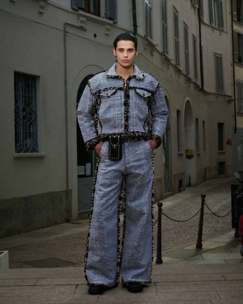 A stylish adult male poses in a light denim jacket and flared pants with patterned accents in an urban setting with grey buildings.