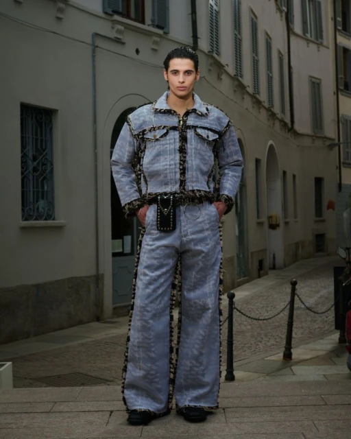 A stylish adult male poses in a light denim jacket and flared pants with patterned accents in an urban setting with grey buildings.