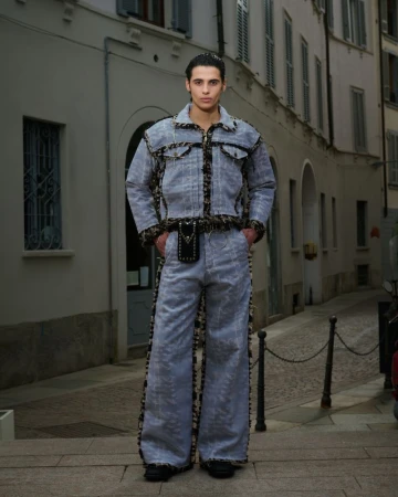 A stylish adult male poses in a light denim jacket and flared pants with patterned accents in an urban setting with grey buildings.