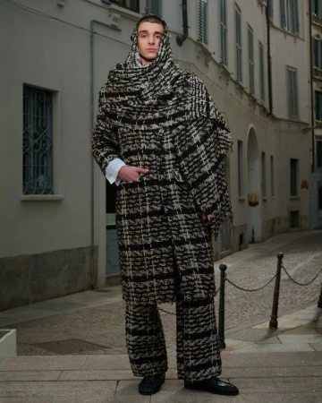Man wearing a textured black and white plaid coat and matching hood standing on an urban street.
