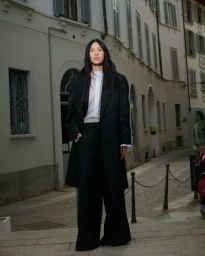 Model with long dark hair wearing a dark coat and trousers standing on an urban street in front of a building facade