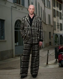 A bald male model wearing a patterned tweed suit standing on a European street with a red moped parked nearby.