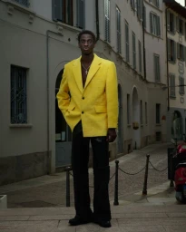 Black man wearing a bright yellow blazer and dark trousers standing on a street in front of a building exterior.
