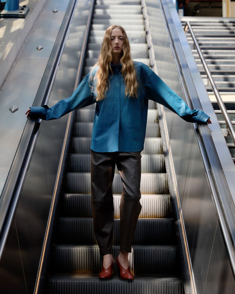 Fashionable adult female posing on an escalator in blue top, brown trousers, and red shoes, creating a stylish and eye-catching composition.