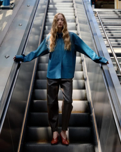 Fashionable adult female posing on an escalator in blue top, brown trousers, and red shoes, creating a stylish and eye-catching composition.