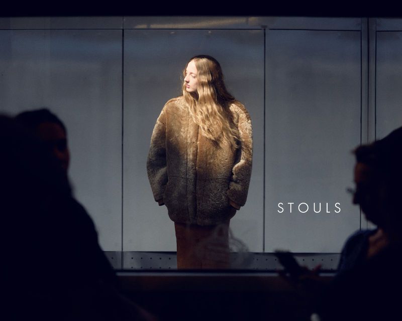 A woman in a furry brown jacket stands in front of a gray background, with two blurred people silhouetted in the foreground.