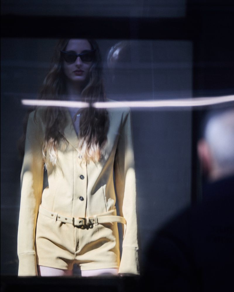 A woman with long hair wears sunglasses and a pale yellow romper in a medium shot, seen through a window with a reflected figure.