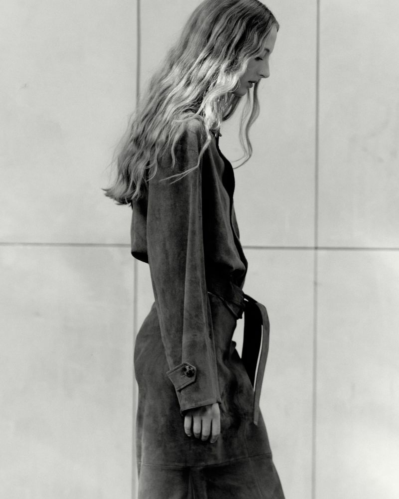 Monochrome profile shot of a blonde woman wearing a long belted coat against a tiled wall, evoking a retro fashion editorial.