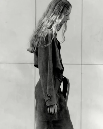Monochrome profile shot of a blonde woman wearing a long belted coat against a tiled wall, evoking a retro fashion editorial.