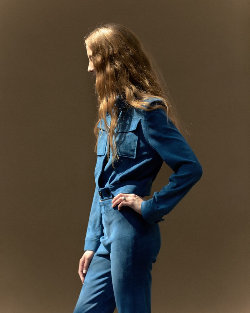 Side profile shot of a woman with long blonde hair wearing a blue pantsuit. The image is shot on a brown background in a photo studio.