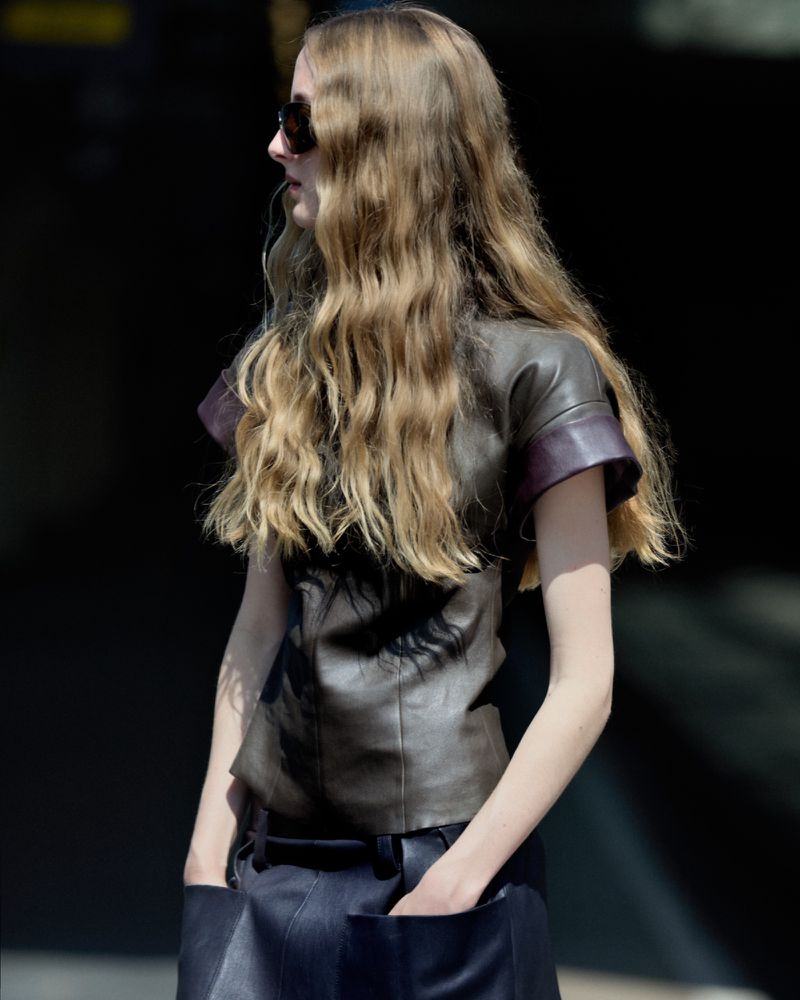 A woman with long wavy blonde hair poses in an outdoor setting, wearing sunglasses, a brown leather top, and a dark skirt. Side view.