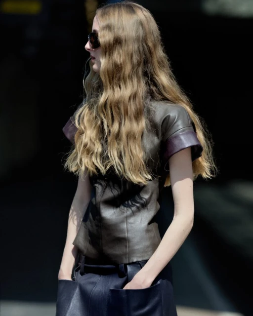 A woman with long wavy blonde hair poses in an outdoor setting, wearing sunglasses, a brown leather top, and a dark skirt. Side view.