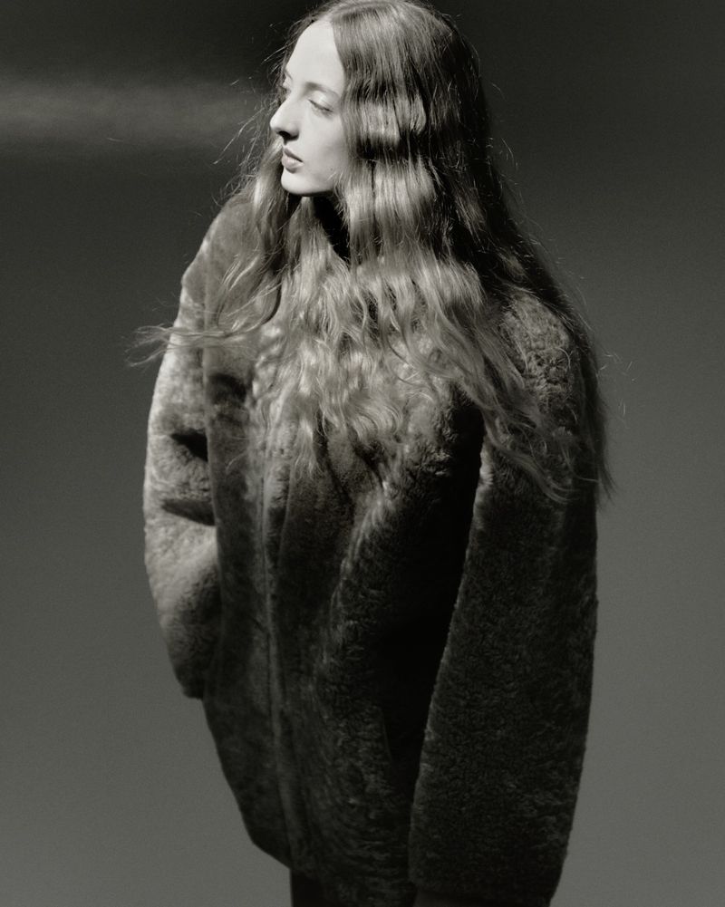 Portrait of a young woman with wavy long hair wearing a textured top, looking away from the camera against a grey background.