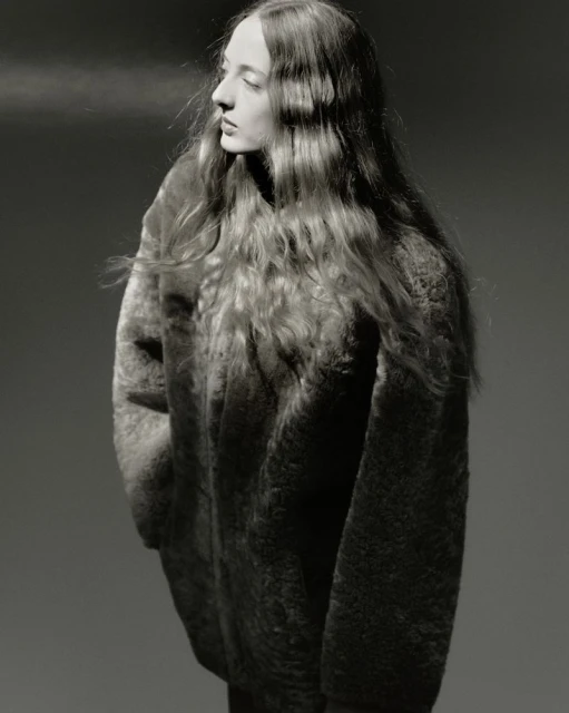 Portrait of a young woman with wavy long hair wearing a textured top, looking away from the camera against a grey background.