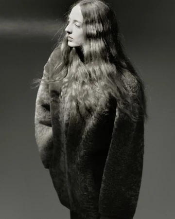 Portrait of a young woman with wavy long hair wearing a textured top, looking away from the camera against a grey background.