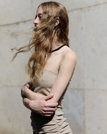 Blonde woman in beige dress poses outdoors with wind blowing hair. Fashion photography features fair skin, neutral colors, natural light.