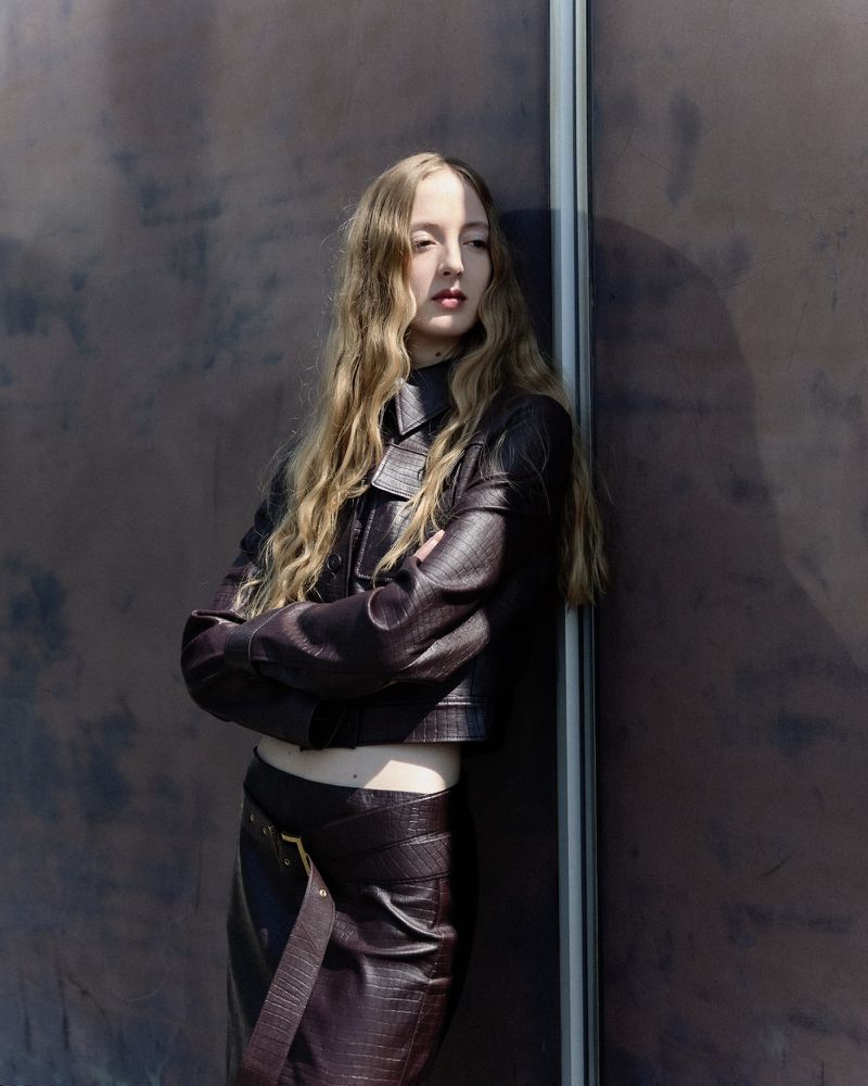 Young adult woman with long blonde hair poses leaning against a wall in a dark brown snakeskin jacket and skirt. Fashion shoot.