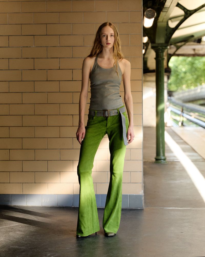 A slender woman poses in a gray halter top and green flare jeans against a brick wall inside a subway station with bright sunlight.