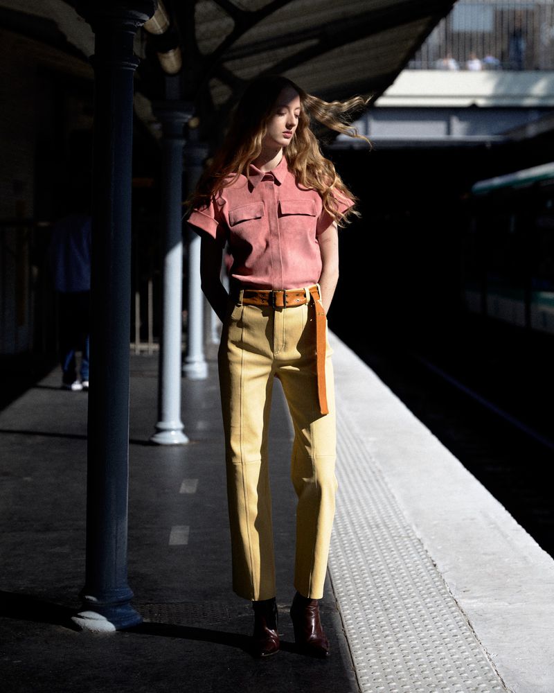 Fashionable woman in pink top and yellow pants stands on a subway platform, bathed in sunlight with shadows from the overhead structure.