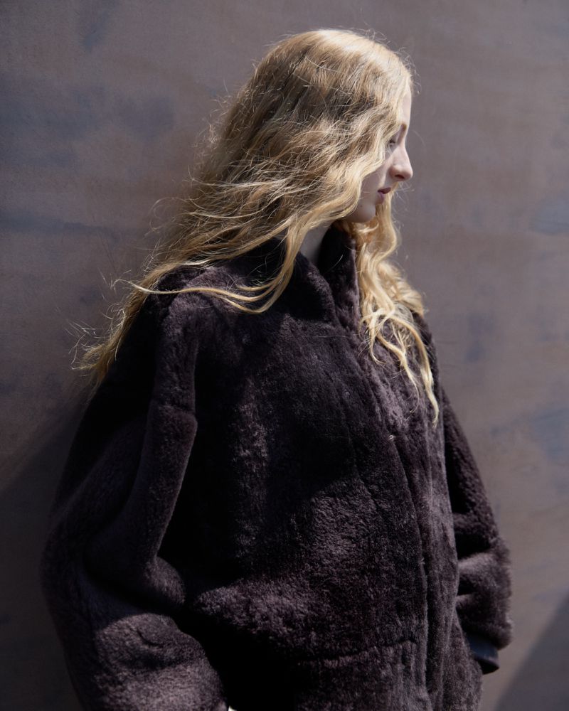Woman with blonde hair wearing a dark brown fur coat, posing in front of a mottled brown and grey background, shot in profile.