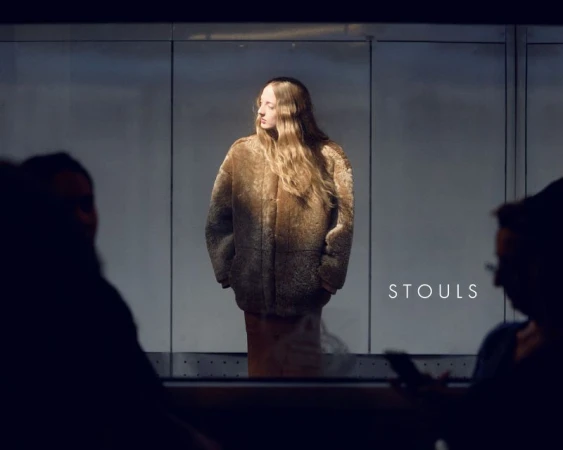 Model in brown shearling jacket standing behind a glass partition with silhouetted figures in the foreground