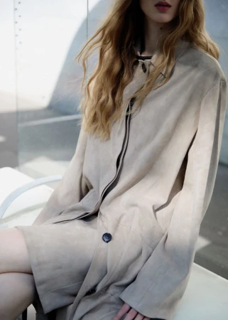Model wearing a light beige coat with long blonde wavy hair against an outdoor architectural wall backdrop.
