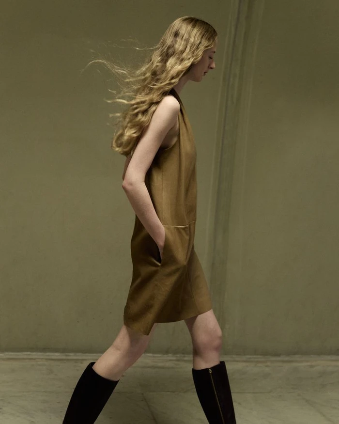 Model with long blonde hair blowing in the wind walking in a brown dress and black boots against a plain wall.