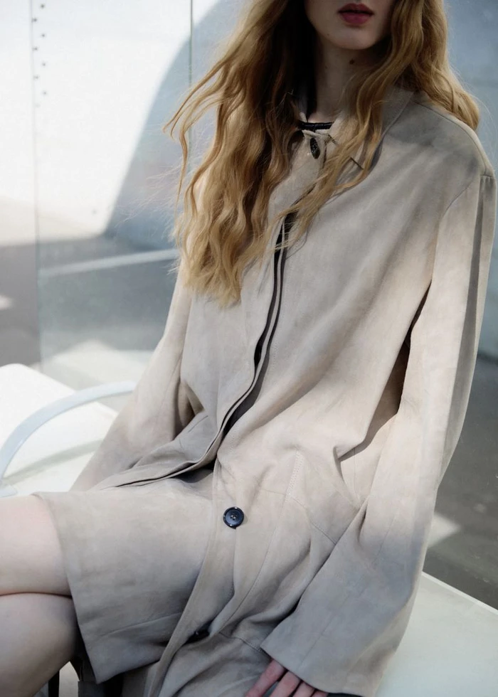 Model wearing a light beige coat with long blonde wavy hair against an outdoor architectural wall backdrop.