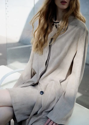 Model wearing a light beige coat with long blonde wavy hair against an outdoor architectural wall backdrop.