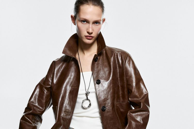 A fair-skinned adult woman with brown jacket poses in a studio shot with a neutral facial expression and simple background. 