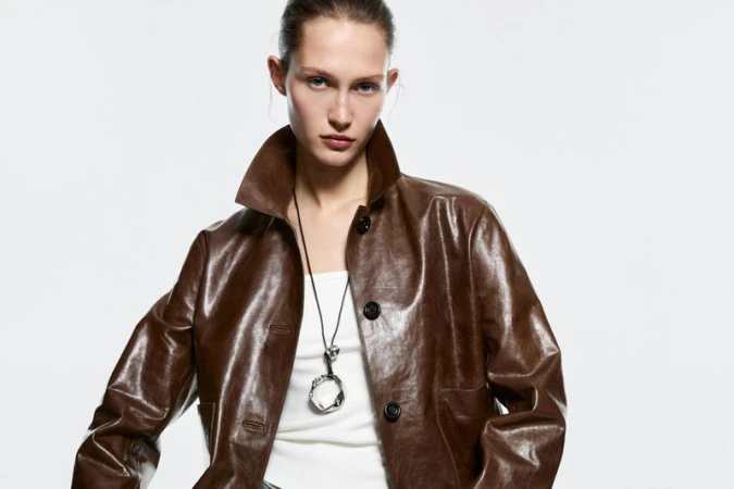 A fair-skinned adult woman with brown jacket poses in a studio shot with a neutral facial expression and simple background. 