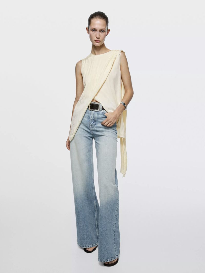 Fashion shot of a woman posing in a sleeveless cream top and light blue jeans in a bright white studio setting, full length portrait.