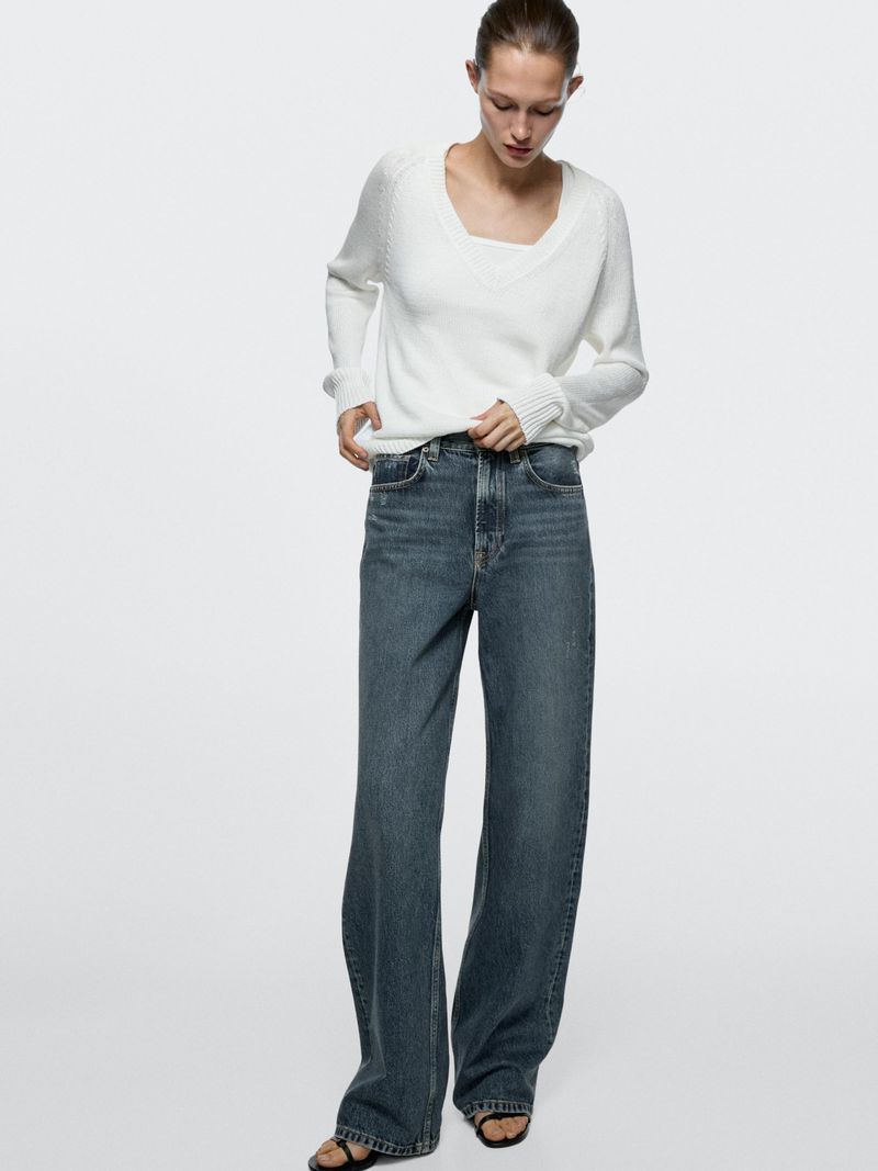 A woman models a white sweater and wide-leg jeans against a neutral background, showcasing a casual yet stylish fashion ensemble.