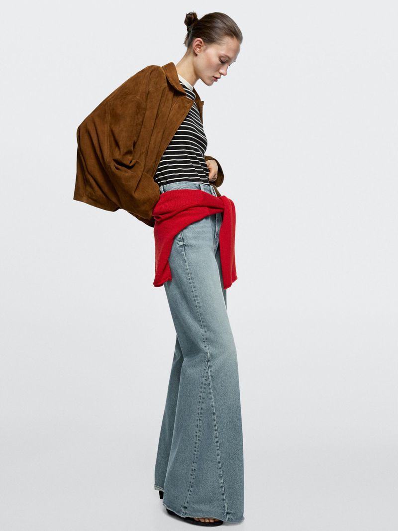 A woman models a brown jacket, striped top, red sweater tied around her waist, and wide-leg jeans in a brightly lit photo studio.